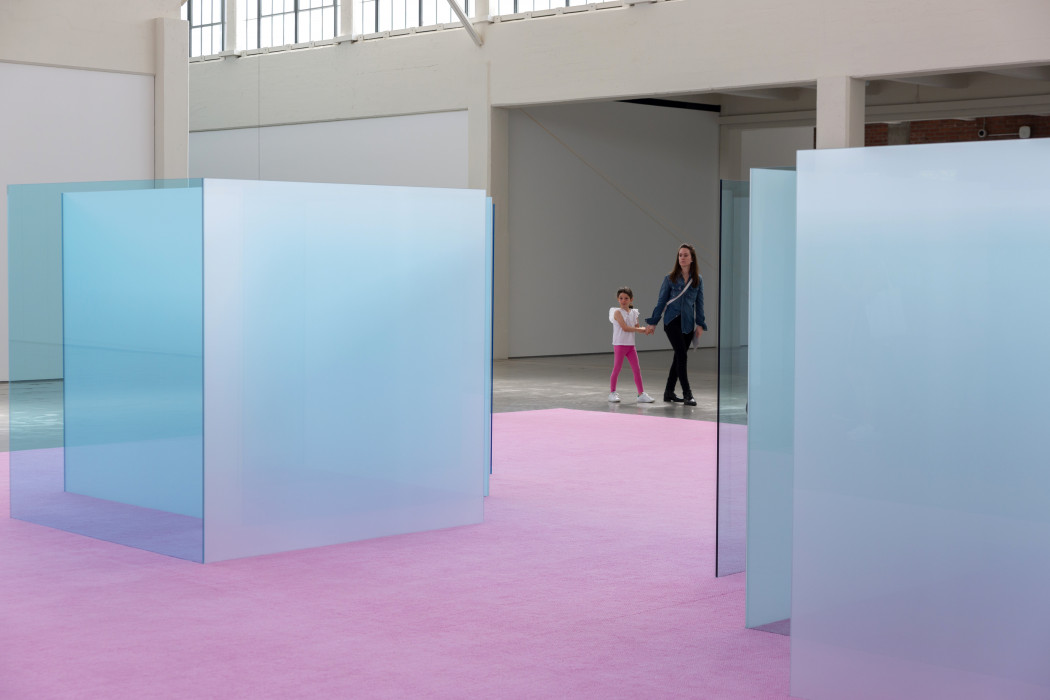 A pink carpet covers the floor and on top of the carpet stands transparent and opaque blue and white glass. A a child and parent hold hands and walk on the concrete floor behind the sculpture, viewable between the two glass panels.